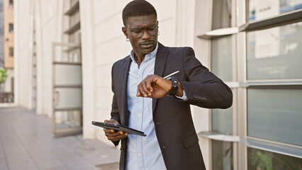 African american businessman checking time outside city building while holding tablet.