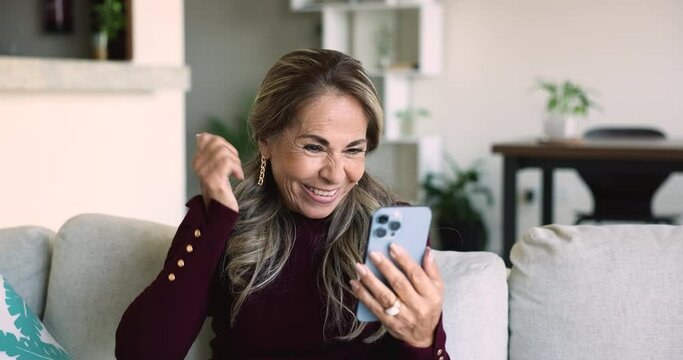 Elderly Mexican woman sit on sofa at home with cellphone, read message, receive great news, get prize reward in web lottery, feels happy, screams, celebrate victory. Success, commercial offer, new app
