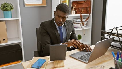 Naklejka premium Focused african man working on laptop in modern office, dressed in business suit.