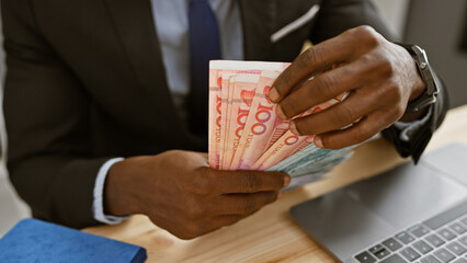 African man in office counts chinese yuan with laptop and notebook in the background