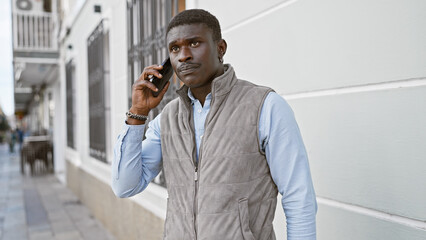 An african man in a vest uses a cellphone on a city street, exuding confidence and style.