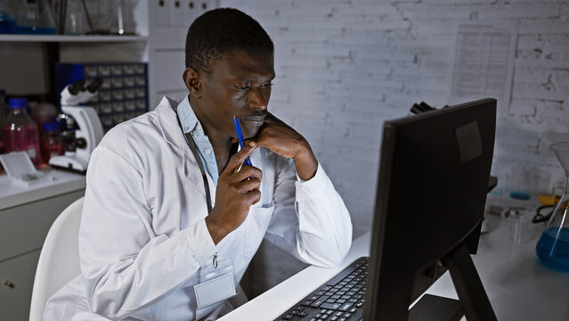 A thoughtful african man in a lab coat analyzes data on a computer in a modern laboratory setting.