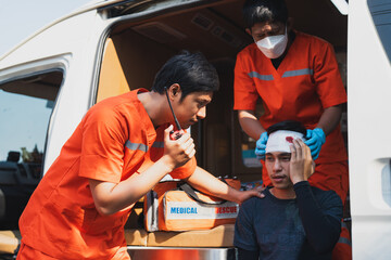 Asian male EMS paramedics or emergency medical technicians in orange uniforms performing first aid applying bandage onto head of young man patient in ambulance from road accident - obrazy, fototapety, plakaty