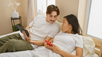 Interracial couple lounging in a bedroom with a man holding a tablet and woman using a smartphone, representing relaxation and connection.