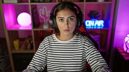 A young hispanic woman with headphones in a colorful gaming room looks surprised at night.