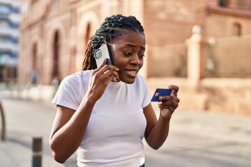 African american woman talking on smartphone holding credit card at street