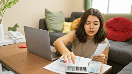 Hispanic woman reviewing finances with calculator, bills, and laptop in home interior.