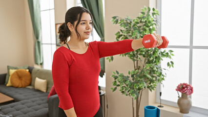 Young hispanic woman exercising with dumbbell in a bright home interior with plants and modern decor.