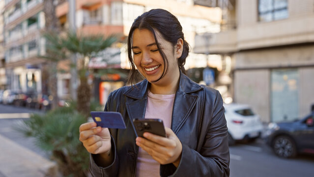 Smiling young hispanic woman holding a credit card and using her smartphone on a sunny urban street.