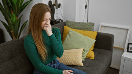 Caucasian woman feeling unwell, sitting on a couch at home with a hand covering her nose, in a cozy living room.