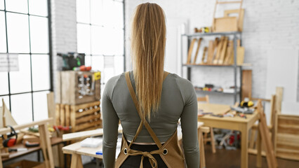 Back view of a young woman with blonde hair standing in a well-lit carpentry workshop, portraying a craftsman atmosphere.