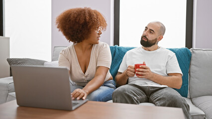 Fototapeta premium Beautiful couple, enjoying a hot cup of coffee, sitting together at home, tightly concentrated on laptop screen while expressing their love.