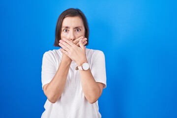 Middle age hispanic woman standing over blue background shocked covering mouth with hands for...
