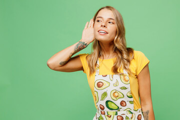 Young curious housewife housekeeper chef cook baker woman wear apron yellow t-shirt try to hear you overhear listening intently isolated on plain pastel green background studio. Cooking food concept.