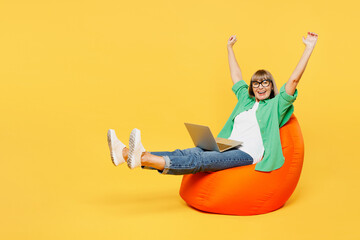Full body elderly IT woman 50s year old in green shirt glasses casual clothes sit in bag chair use work on laptop pc computer do winner gesture isolated on plain yellow background. Lifestyle concept