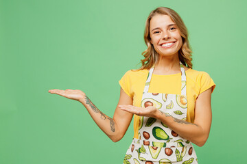 Young smiling happy cheerful housewife housekeeper chef cook baker woman wear apron yellow t-shirt point hands arms aside on area isolated on plain pastel green background studio Cooking food concept