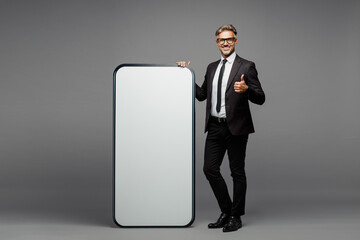 Full body fun adult employee business man corporate lawyer wear formal black suit shirt tie work in office big huge blank screen area mobile cell phone show thumb up isolated on plain grey background.