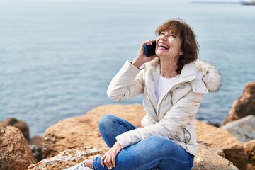 Middle age woman talking on smartphone sitting on the rock at seaside