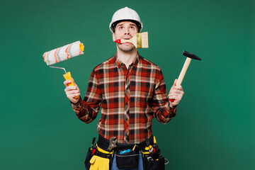 Young employee man wears red shirt hardhat hat hold paint roller hammer brush in mouth isolated on...