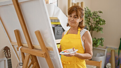 A mature woman artist in a yellow apron paints in a well-lit art studio, holding a palette and brush.