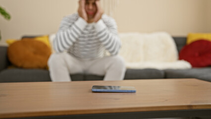 Worried young hispanic man waits nervously for a smartphone call, sitting home on the sofa, a...