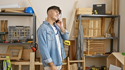 Smiling young hispanic carpenter confidently talking business on smartphone in bustling carpentry workshop