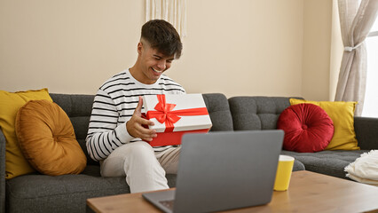 Surprised young hispanic man sitting at home on the sofa, unpacking surprise gift while happily speaking and smiling during online video call. - obrazy, fototapety, plakaty