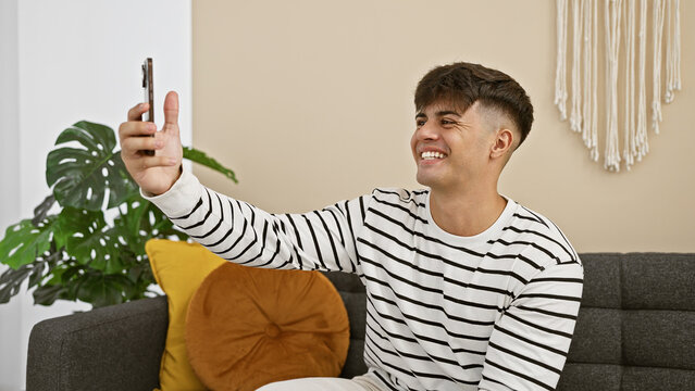 Cheerful young hispanic man joyfully capturing a selfie with smartphone while chilling on the sofa in home's comfortable living room.