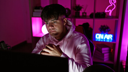 Focused young hispanic man streamer, sitting in his dark gaming room, fully invested in the virtual...