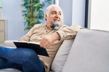 Middle age grey-haired man reading book sitting on sofa at home