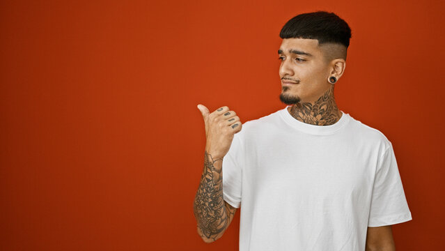 Cool tattooed latin man, in a casual style, seriously pointing to the side, over a stark, isolated red background