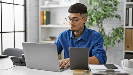 Concentrated young latin man at work, elegantly dressed, business professional operating a laptop and touchpad in his office environment. a serious, focused worker managing success, indoors.