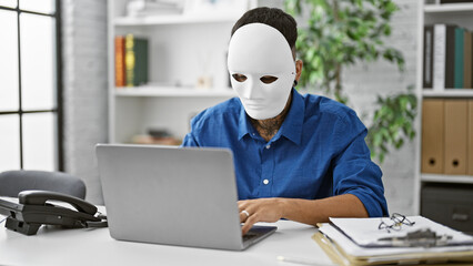 Masked latin hacker, young man unleashes a daring cyber attack on office data security with his laptop in a tattooed, focused act of online crime