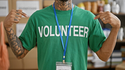 Handsome young latin man, tattooed and working, selflessly pointing at his volunteer uniform inside...