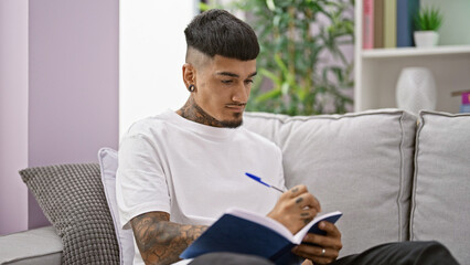 Handsome young latin man sitting on his living room sofa, engrossed in writing on a notebook, ideas...