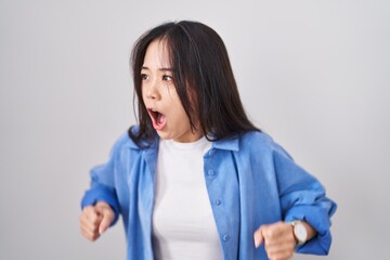 Young chinese woman standing over white background angry and mad screaming frustrated and furious,...