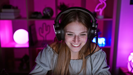 A smiling young woman with headphones enjoys gaming in a vibrant purple-lit room at night.