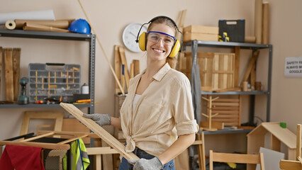 Smiling woman with safety gear holding lumber in a carpentry workshop