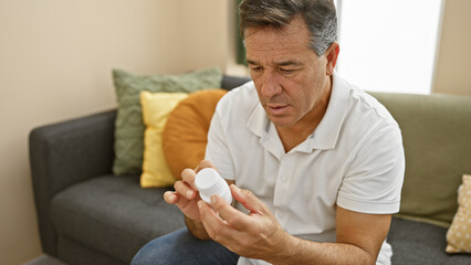 Mature man sitting on couch at home, examining a pill bottle thoughtfully.