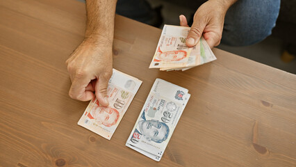 Man counting singaporean dollars at a wooden table indoors, portraying finance and domestic setting.