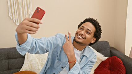 Smiling man taking selfie on couch indoors with red phone, showing peace sign and casual style.