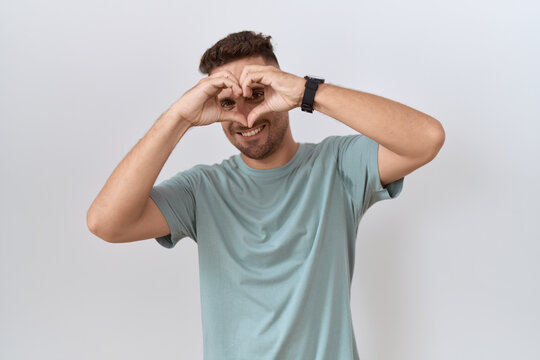 Hispanic man with beard standing over white background doing heart shape with hand and fingers smiling looking through sign