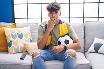 Hispanic teenager sitting on the sofa watching football match covering mouth with hand, shocked and...