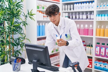 Young hispanic teenager pharmacist using computer holding pills bottle at pharmacy