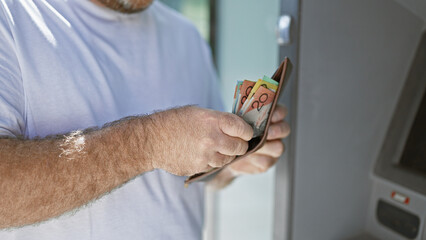 Handsome middle-aged caucasian man outside on urban street, captivating portrait of him holding wallet counting australian cash dollars at atm machine, engaging in banking transaction. © Krakenimages.com