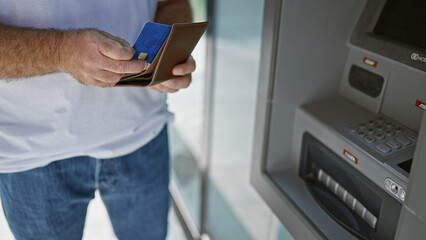 Handsome middle-aged caucasian man making a credit card transaction at a street atm. captivating portrait of an attractive adult male handling personal finances in an urban exterior. - obrazy, fototapety, plakaty