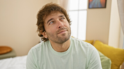 Fototapeta premium Thoughtful young man with beard looking up in a cozy bedroom setting, embodying casual home life.
