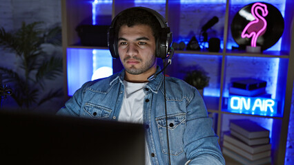A young man wearing headphones sits at a desk in a dark, neon-lit gaming room, looking intently at a computer screen.