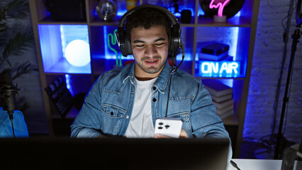 A young man with headphones in a dark gaming room looks at a smartphone, highlighting technology...