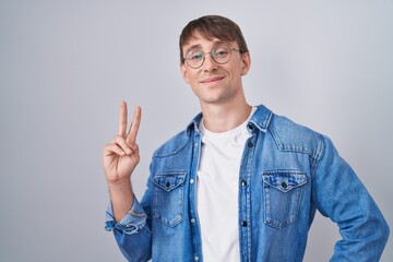 Caucasian blond man standing wearing glasses smiling looking to the camera showing fingers doing victory sign. number two.